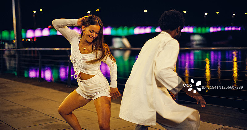 年轻情侣在河边大道上表演现代快节奏舞蹈，笑容满面，水面倒映灯光图片素材