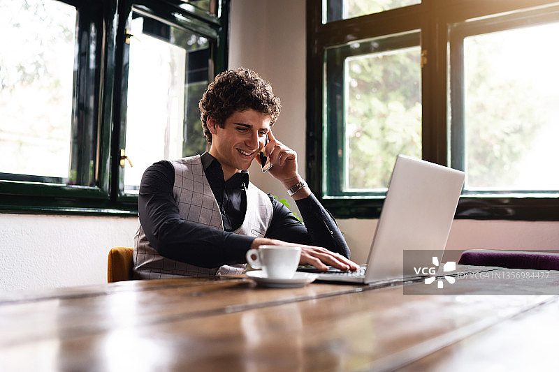 使用笔记本电脑并通过手机交谈的现代快乐的年轻商人图片素材