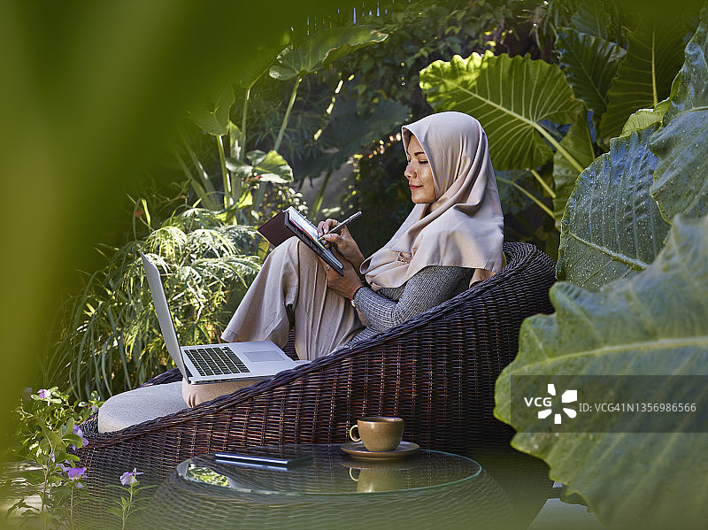 戴着头巾的亚洲女人在花园家具旁工作，周围环绕着热带植物，在笔记本上写作图片素材