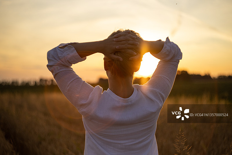 穿着白衬衫、举起双臂站在日落时小麦地里的女人图片素材