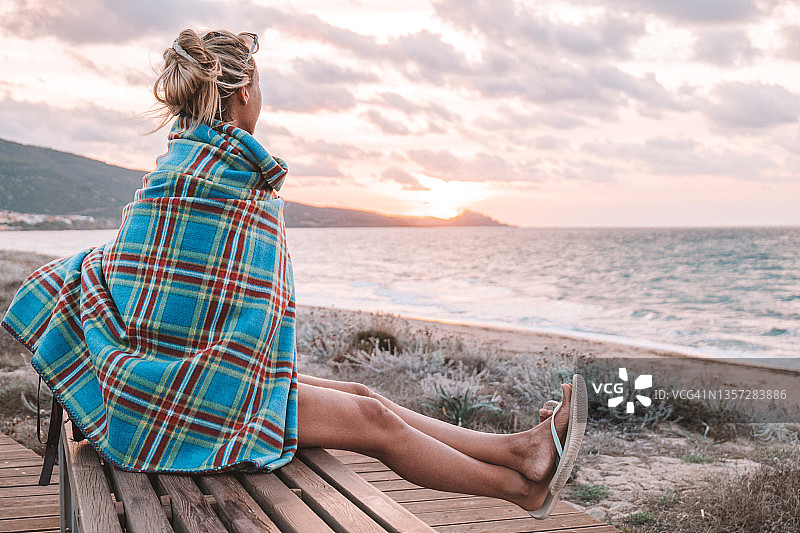 快乐的年轻女人裹着毯子，在木制海滩的木板路上观看日落图片素材