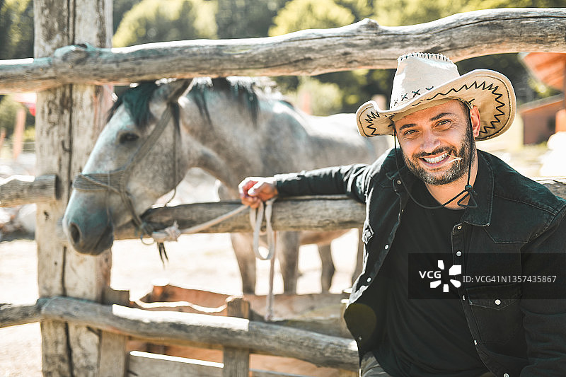 快乐的乡村男牛仔嘴里叼着稻草坐在拴在牧场的马旁边图片素材