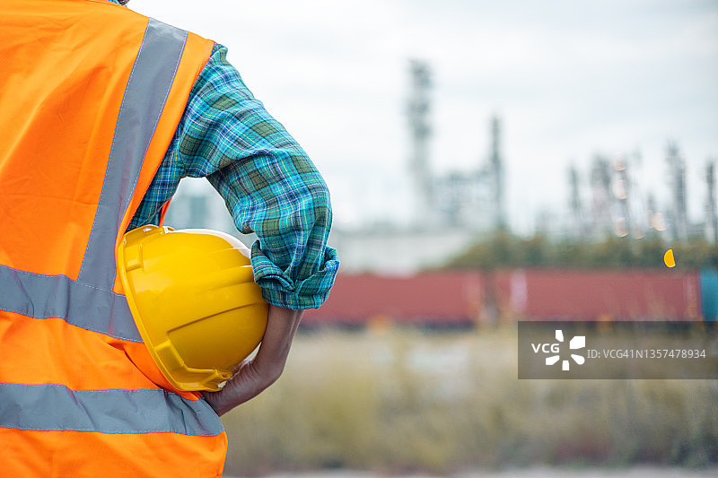 身穿西装，手持白色安全帽的工程师背影，背景为炼油厂图片素材