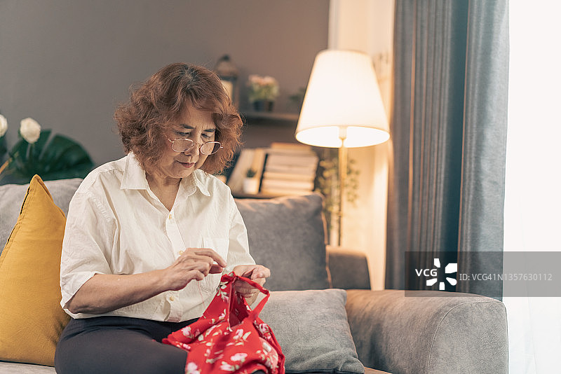 年长的亚洲妇女在家中客厅刺绣，为女儿缝制衣服。退休后的老妇人在空闲时享受自己的爱好并微笑，穿着休闲图片素材