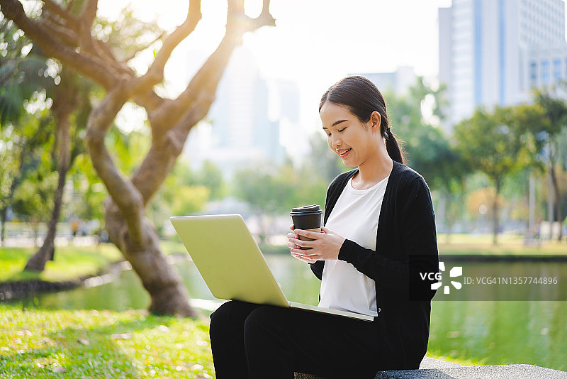 在现代城市公园里户外使用笔记本电脑喝咖啡的年轻亚洲女商人图片素材
