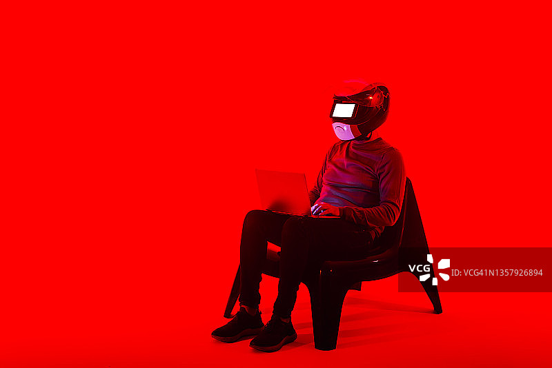 戴着未来主义头盔在红色背景下使用笔记本电脑的男人图片素材