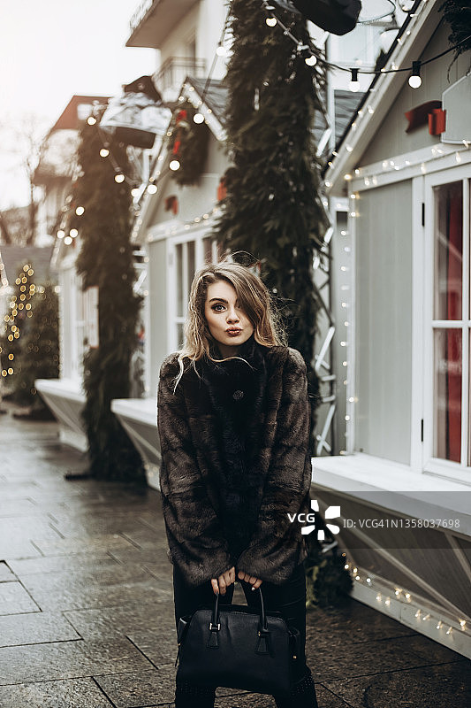 穿着皮草大衣的年轻女子站在圣诞装饰的小屋前图片素材