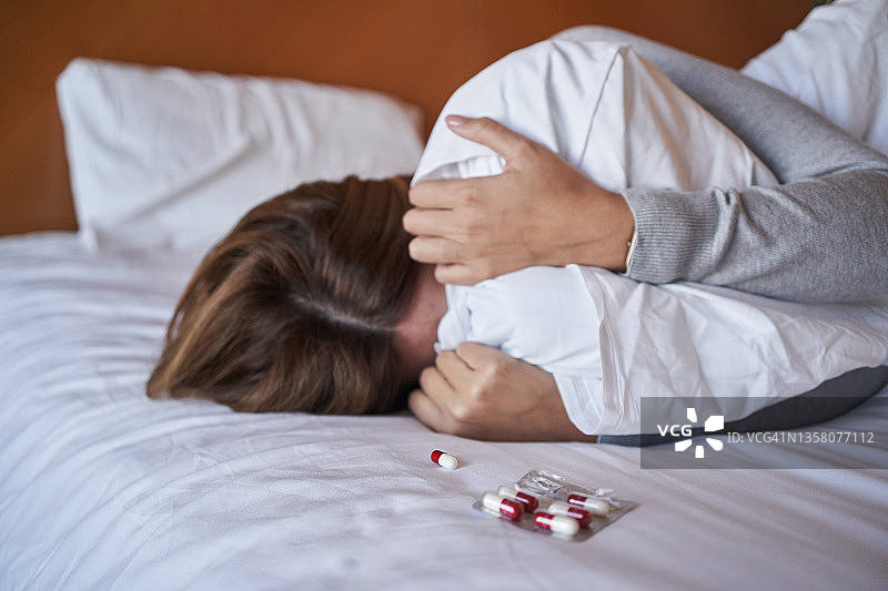 年轻女人哭泣时用枕头遮住脸，旁边放着药片图片素材