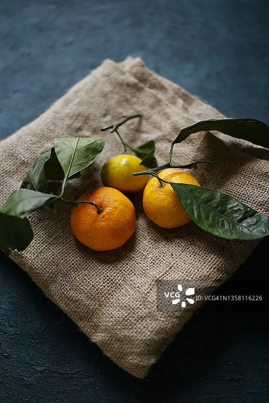 深蓝绿色粗糙水泥地面上，粗麻布上的三个带叶成熟橘子图片素材