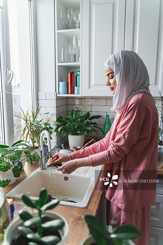 年轻的穆斯林中东妇女在白色现代厨房的水槽旁洗碗图片素材