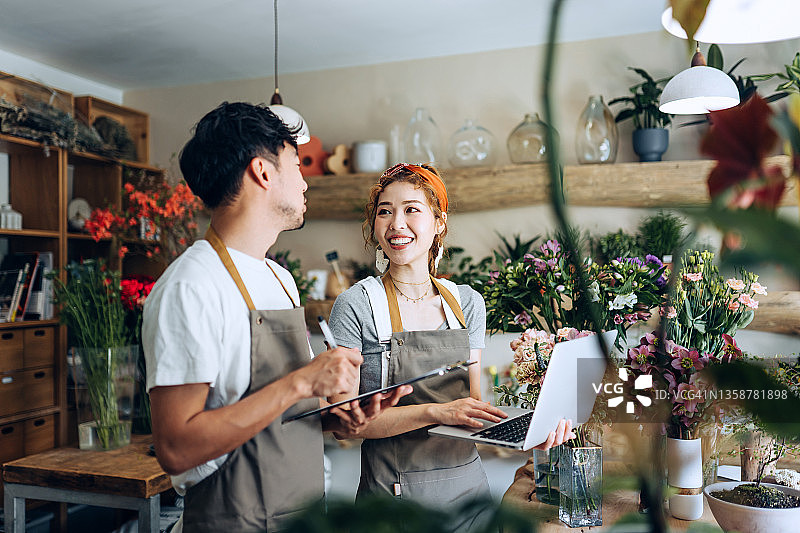 年轻的亚洲夫妇在花店使用笔记本电脑一起工作，讨论业务，鲜花植物背景。创业、商业合作和团队合作，共同努力实现业务成功图片素材
