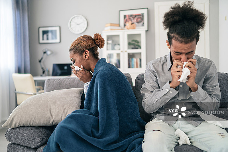 生病的年轻非裔美国夫妇坐在家里的沙发上，两人都在擤鼻涕，女人身上盖着毯子图片素材
