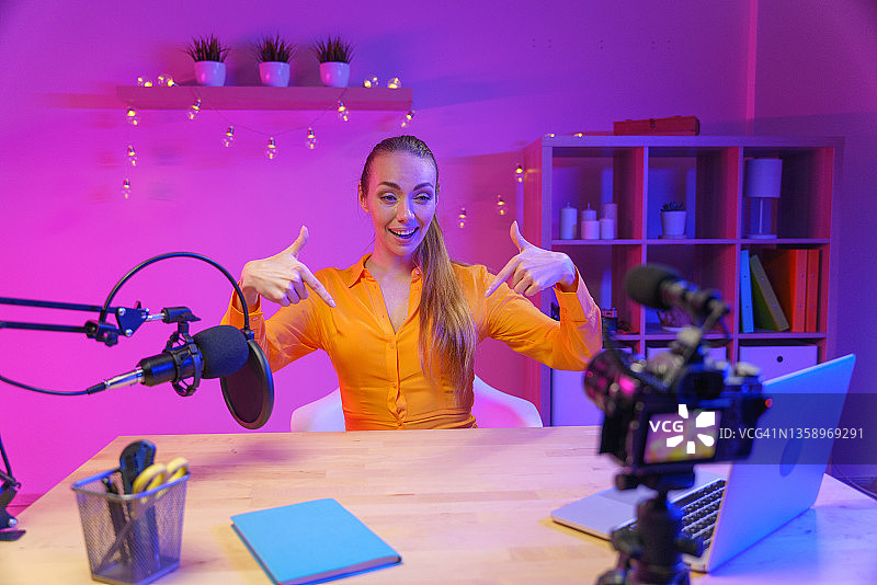 一位穿着黄衬衫的金发年轻迷人博主，面带微笑地指向拍摄相机中的链接位置图片素材