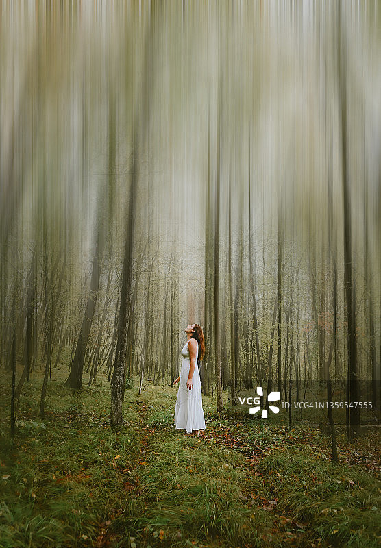 身穿白色连衣裙的女人独自站在模糊的树林空地上图片素材