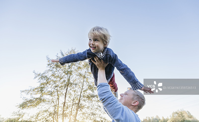 微笑的男人高举着金发男孩，在晴朗的天空下飞翔图片素材