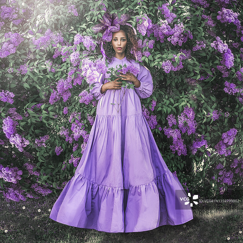 身穿紫色长裙、头戴头饰的非洲年轻女子在丁香树丛旁图片素材