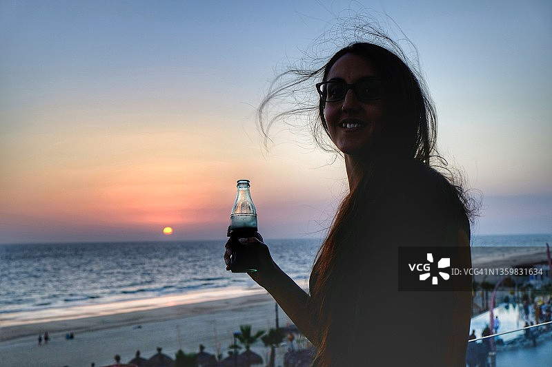 日落时手持饮品、背景为红色太阳的女人剪影图片素材