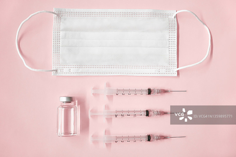 戴着口罩并接种疫苗：粉色背景上的三个带针头的注射器、医用小瓶和口罩图片素材