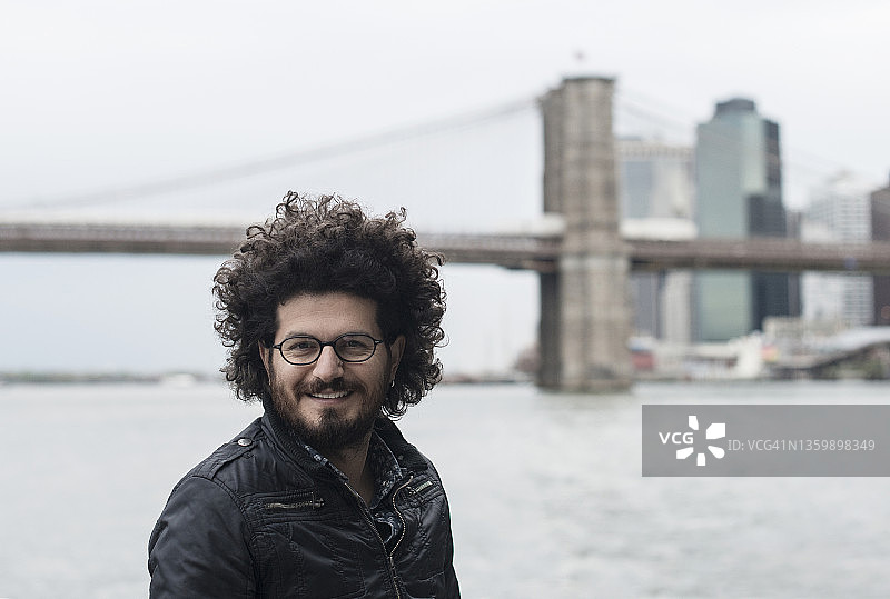 在纽约市的卷发年轻男子肖像。背景可见布鲁克林大桥图片素材