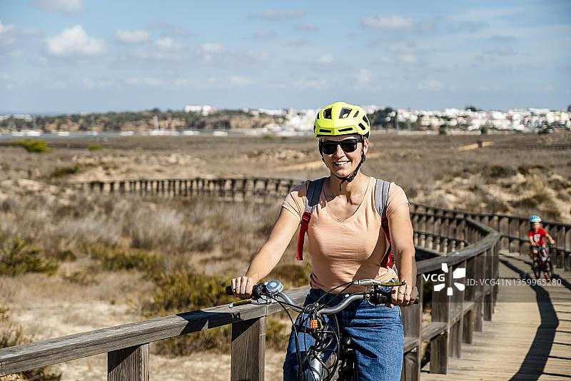 母亲在葡萄牙阿尔加维阿尔沃尔的木制走道上骑自行车，儿子在车上图片素材