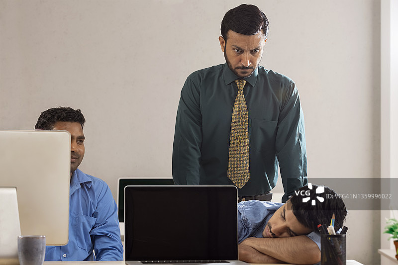 生气的上司在两名公司员工后，其中一人在办公桌上睡觉。图片素材