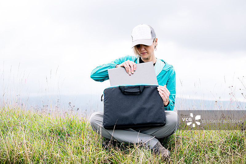 女人把笔记本电脑放进包里，坐在山坡的草地上图片素材