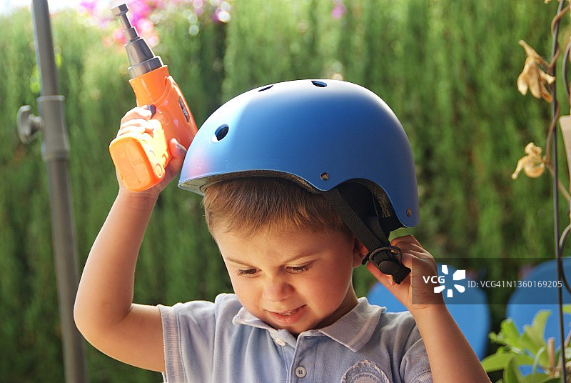 戴着自行车头盔，手持玩具钻头的愤怒男孩站在花园里图片素材