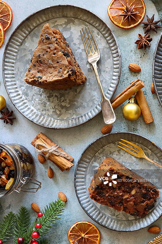 自制圣诞水果蛋糕切片图片：盘子、叉子、肉馅蜜饯罐、金色小饰品、肉桂棒、杏仁、柑橘干果、红莓、云杉针、八角茴香，高角度视图图片素材