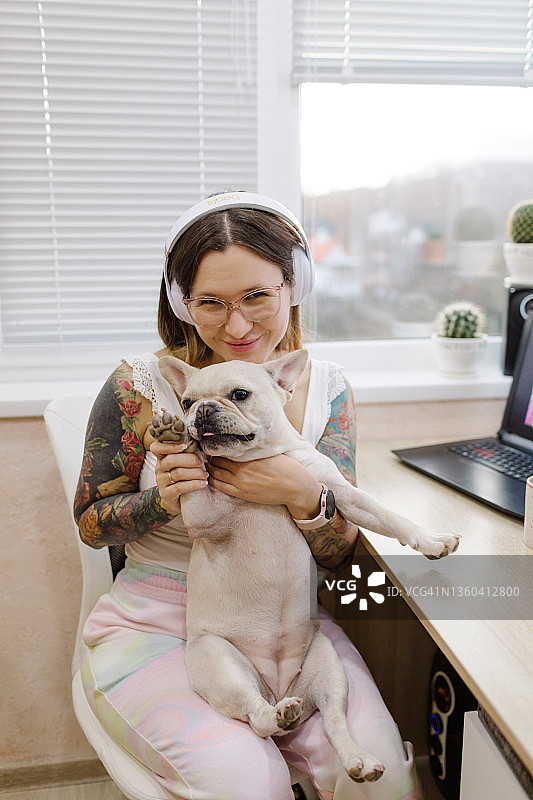 戴着眼镜和耳机长发的年轻欧洲女人抱着白色法国斗牛犬坐在她的手臂上，通过笔记本电脑进行视频通话图片素材