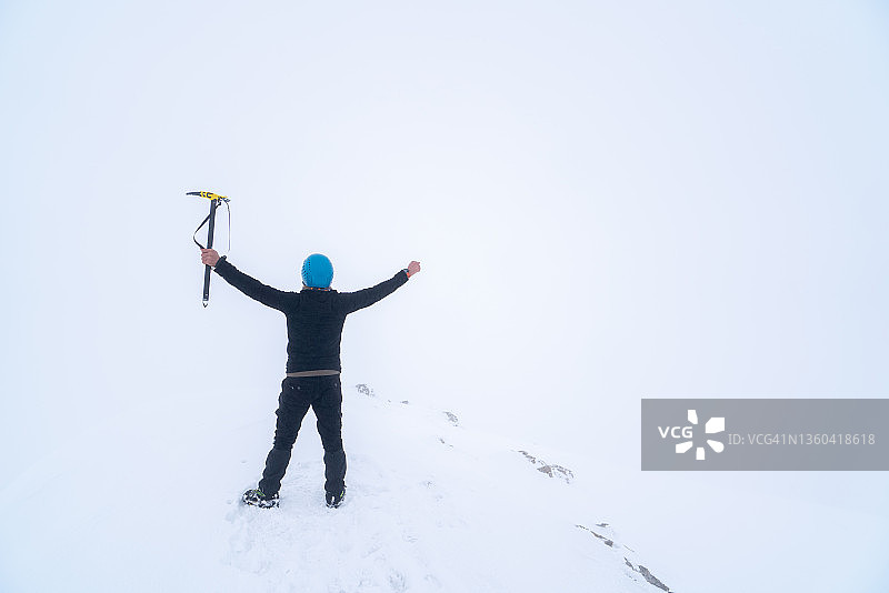 成功的年轻登山者在雾蒙蒙的冬季天气里举起手臂，在高海拔雪山的顶峰握着冰镐图片素材