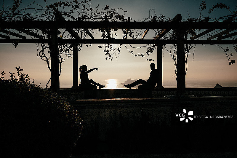 情侣在里斯本圣卢西亚观景台栏杆边相对而坐，俯瞰大海的剪影图片素材