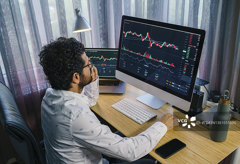 商人在家分析电脑和笔记本电脑屏幕上的加密货币市场图表。比特币/加密货币概念图片素材