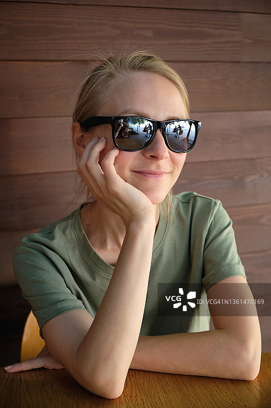 戴太阳镜的时尚女孩坐在户外咖啡馆或餐厅的木桌旁，等待她的订单，眺望远方。美味的午餐，小吃，快餐。快乐微笑的年轻女人肖像图片素材