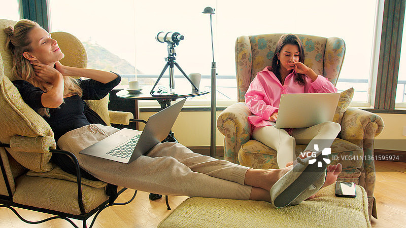 女同事在全景窗边使用笔记本电脑喝咖啡，欣赏壮丽海景，工作期间聊天欢笑图片素材