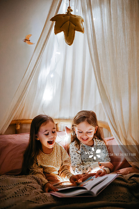 两姐妹坐在床上，笑着阅读一本有趣的读物图片素材