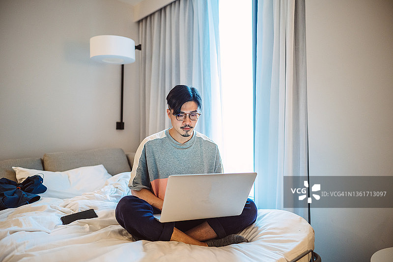 疫情隔离期间，在酒店房间里用笔记本电脑工作的年轻英俊男人图片素材