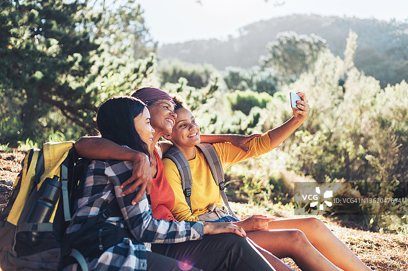 快乐的母亲和女儿们在阳光明媚的树林里徒步旅行自拍图片素材