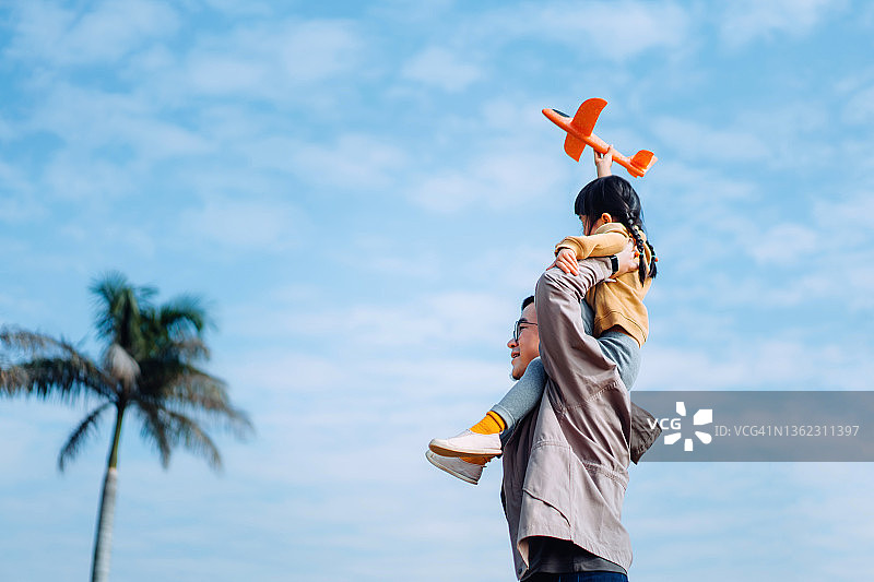 快乐的年轻亚洲父亲肩扛女儿，在阳光明媚的公园里一起玩耍飞机玩具图片素材