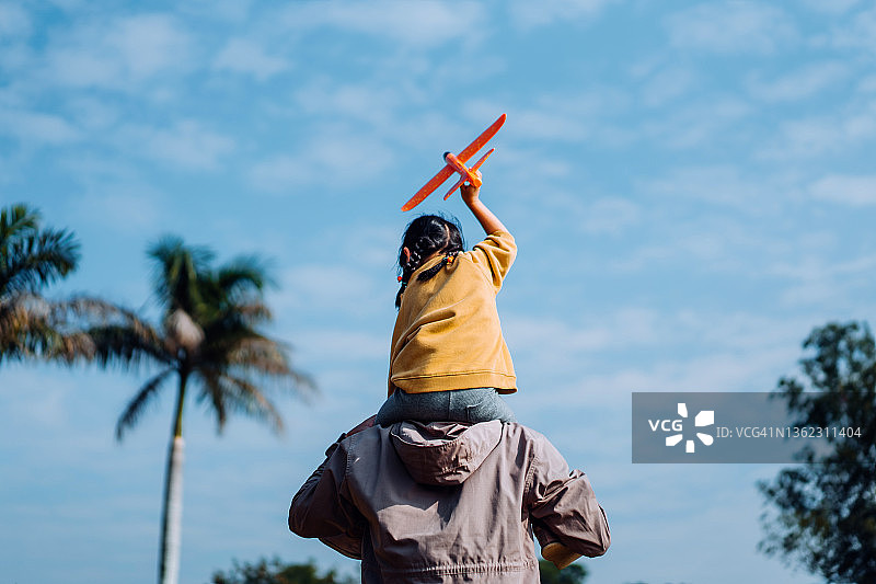 快乐的年轻亚洲父亲肩扛女儿，在阳光明媚的公园里一起玩耍飞机玩具图片素材
