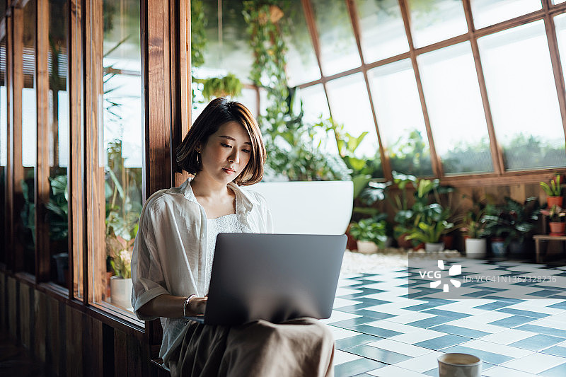 年轻亚裔女性在家使用笔记本电脑工作，坐在玻璃窗框边的地板上，远程办公、自由职业者、小企业概念图片素材