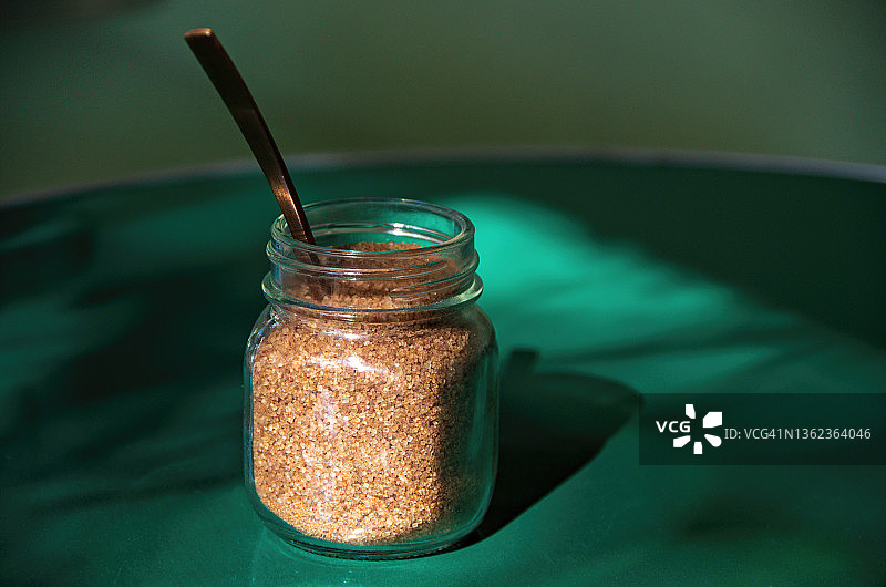 小罐特比纳多糖（原糖）和黄铜茶匙，在绿色人行道咖啡桌上图片素材