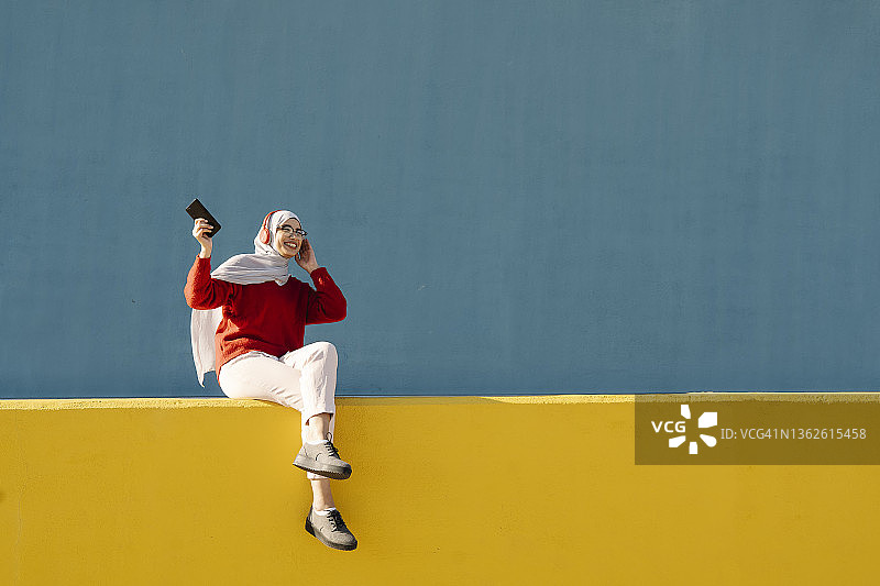 年轻的穆斯林妇女在户外用智能手机和耳机听音乐。图片素材