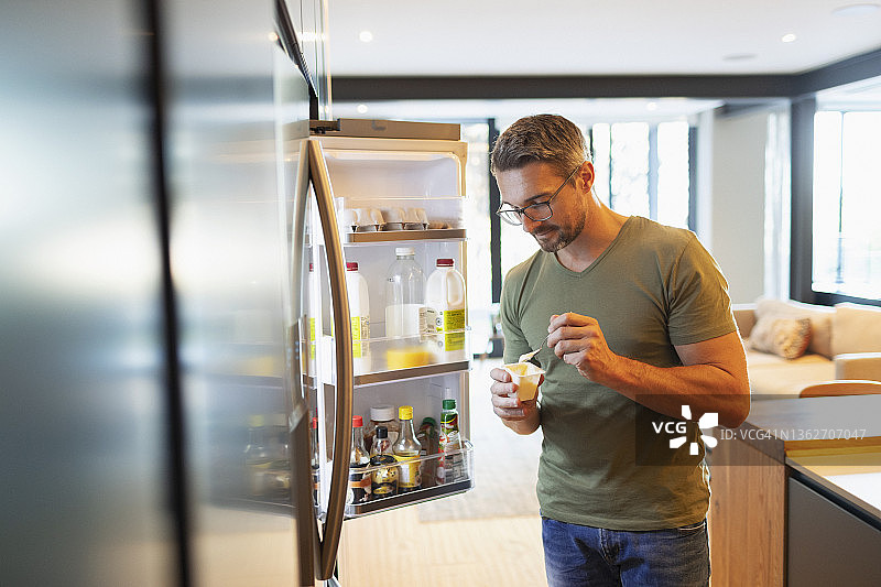男人在厨房的开放式冰箱旁吃酸奶图片素材