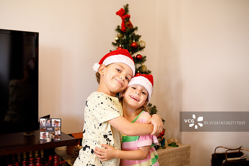 戴圣诞帽的年幼兄妹在圣诞树背景下拥抱并看向镜头图片素材