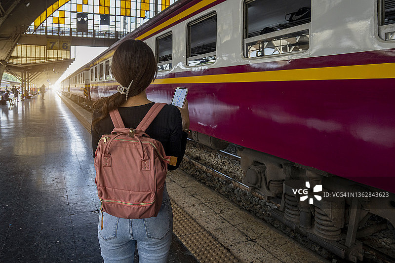 年轻女子游客，带着天蓝色背包和帽子，在曼谷火车站的火车背景下看着地图。在泰国曼谷旅行。旅游概念图片素材