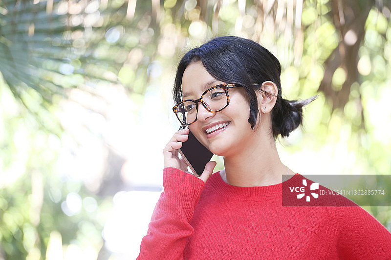 一位拉丁裔千禧一代女子，深色头发盘成发髻，站在花园里一边工作一边用手机通话，身穿红色毛衣和眼镜。图片素材
