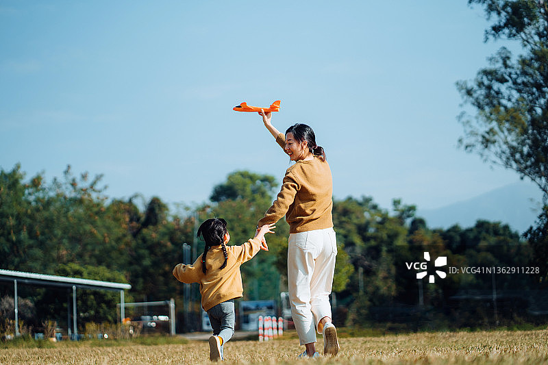 年轻的亚洲母亲和可爱的小女儿在户外共度时光，在美丽的蓝天下，在公园里玩飞机玩具，笑容满面图片素材