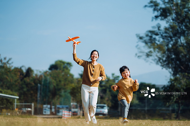 年轻的亚洲母亲和可爱的小女儿在户外共度时光，在美丽的蓝天下，在公园里玩飞机玩具，笑容满面图片素材