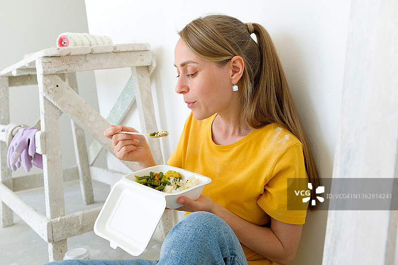 一位快乐的年轻女子或艺术家坐在彩绘墙边吃东西。一名建筑工人在建筑工地的地板上盘腿而坐。家居和专业维修与施工。建筑行业，午休和小吃。图片素材