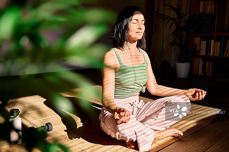女人在家练习瑜伽和冥想，以莲花坐姿坐在瑜伽垫上，闭着眼睛放松。正念冥想概念。健康。图片素材
